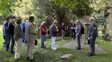 Das Foto zeigt eine Gruppe von Menschen, die nach der Gedenkveranstaltung noch an einer Führung zu den Kriegsgräbern auf dem Friedhof teilgenommen haben. Die Gruppe wird durch Jürgen Pohl und Georg Möllers vom Verein für Orts- und Heimatkunde geführt. Auf dem Foto erläutert Jürgen Pohl der Gruppe ein besonderes Denkmal auf dem Friedhof.